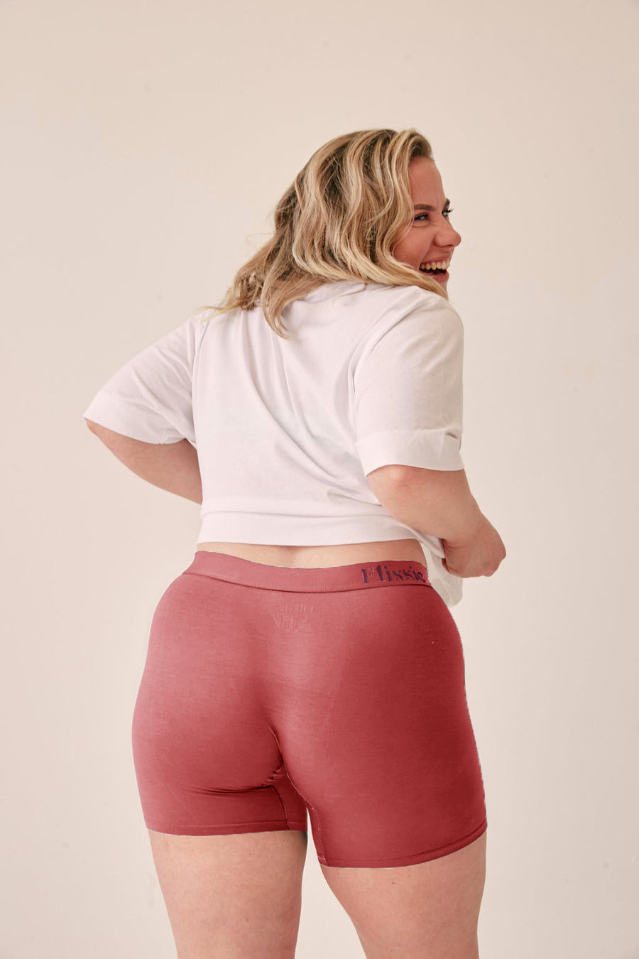 Model in Flissie women's bamboo boxer shorts in plum pink and a white T-shirt, smiling over her shoulder